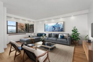Modern living room with a large gray sofa, two wooden armchairs, glass coffee table, abstract wall art, and a large window overlooking trees.