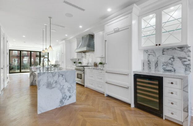 Bright modern kitchen with white cabinetry, marble countertops, a large marble island, stainless steel stove, and wood herringbone floor.