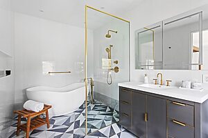 Modern bathroom with a freestanding bathtub, glass-enclosed shower with gold fixtures, and dark vanity with dual sinks under a large mirror.