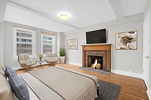 Modern bedroom with a bed, two chairs by large windows, a brick fireplace with a fire, and a wall-mounted TV above it.