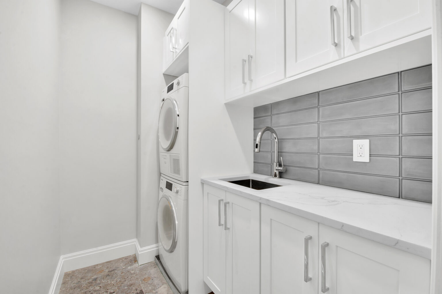 Modern laundry room with stacked white washer and dryer, white cabinetry, marble countertop, stainless steel sink, and gray subway tile backsplash.
