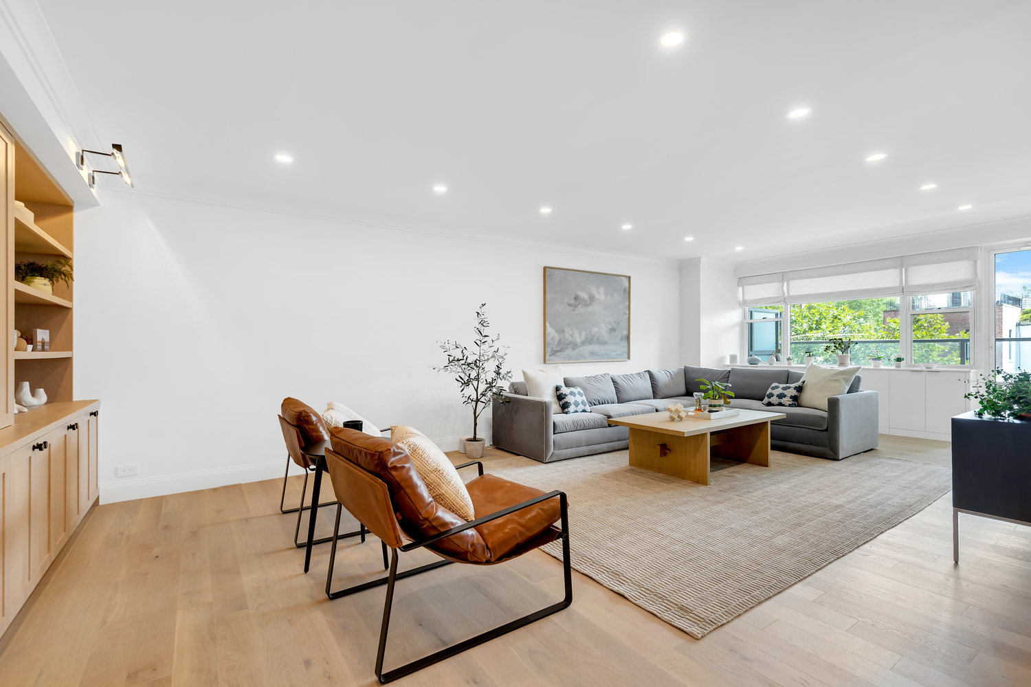 Bright modern living room with light wood flooring, gray sectional sofa, brown leather chairs, large rug, and a wooden coffee table under recessed ceiling lights.