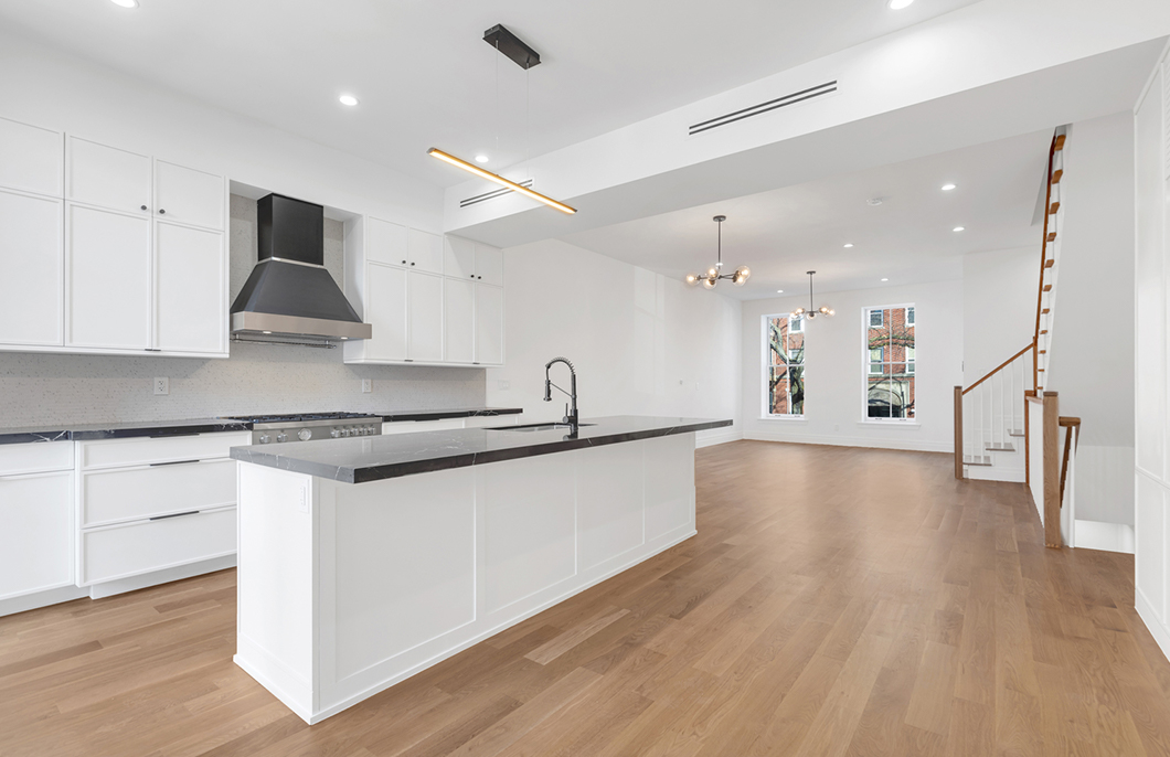Modern open-plan kitchen and living area with white cabinetry, black countertops, wood flooring, and two pendant lights near windows.