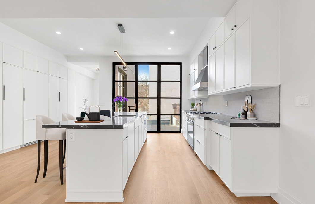 Modern bright kitchen with white cabinets, black marble countertops, wooden floor, an island with bar stools, and large glass door at the end.