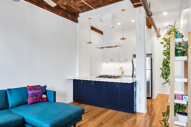 Modern kitchen with white upper cabinets, navy blue lower cabinets, gold faucet, stainless steel refrigerator, and a turquoise sectional sofa with colorful pillows on wooden flooring.