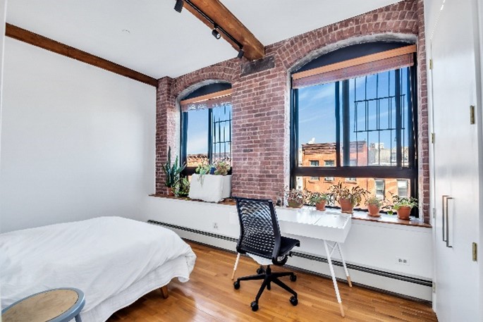 Bright bedroom with exposed brick walls, wooden ceiling beam, two large windows with plants on the sill, a white bed, and a white desk with a black swivel chair.