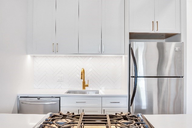 Modern white kitchen with stainless steel refrigerator, gold faucet, gas stove, and white cabinetry with herringbone tile backsplash.