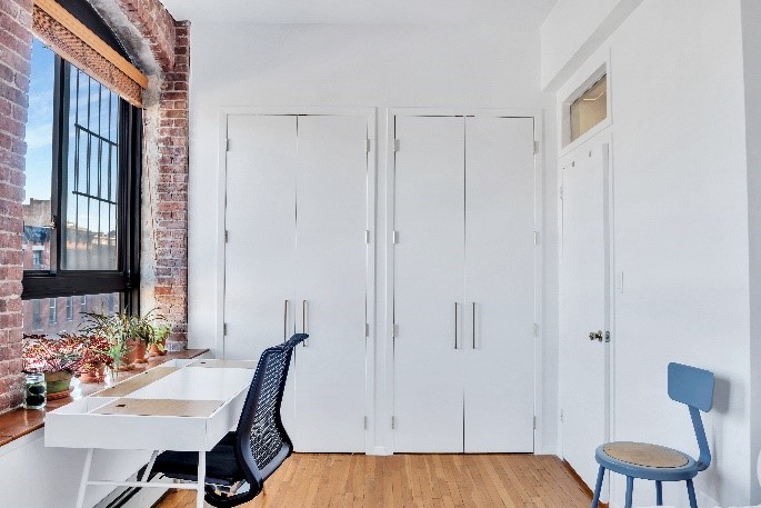 Minimalist room with wooden floor, white desk under window with potted plants, black office chair, and white closet doors.