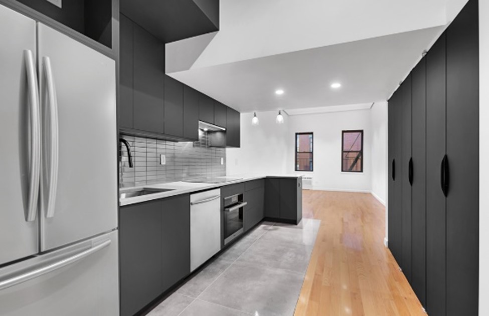 Modern kitchen with black cabinets, stainless steel refrigerator, oven, dishwasher, gray tiled floor, and wood flooring transitioning to a dining area with two windows and closet doors on the right.