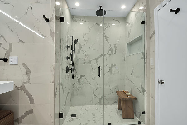 Modern shower with glass doors, rain showerhead, handheld shower, marble-patterned tiles, and a wooden bench.