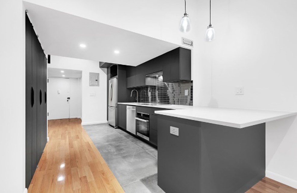 Modern kitchen with black cabinets, stainless steel appliances, gray tiled floor, white countertop, and wooden flooring in the adjacent area.