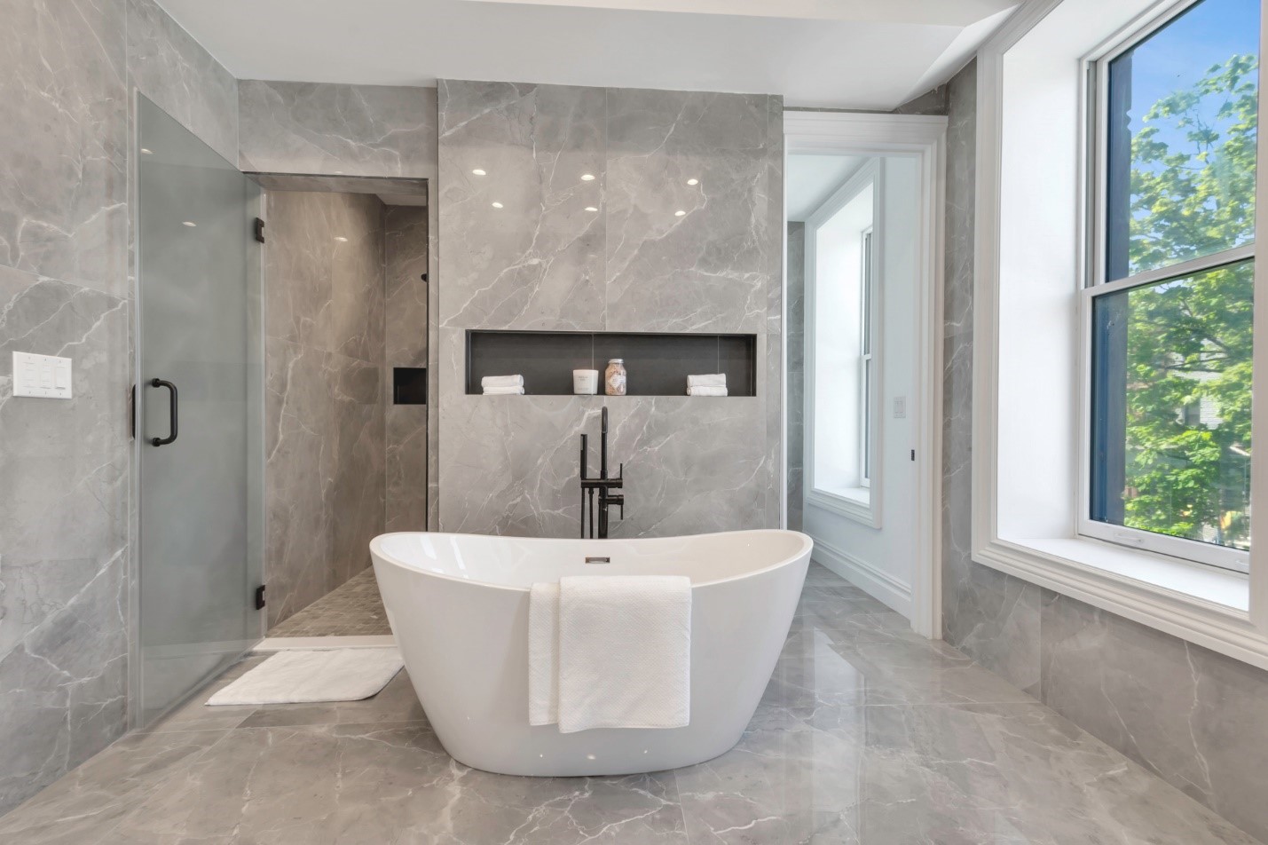 Modern bathroom with freestanding white bathtub, gray marble walls and floor, recessed shelf with towels and decor, and large window.