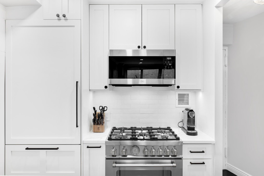 Modern white kitchen with stainless steel gas stove, overhead microwave, knife block, and coffee machine.