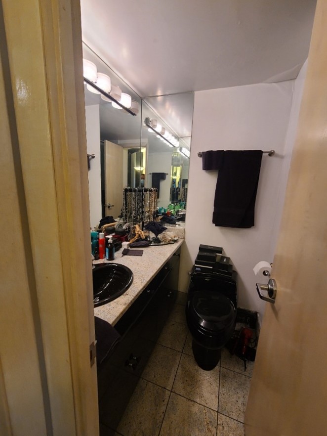 Small bathroom with black sink and toilet, beige countertop cluttered with toiletries, and black towel hanging on wall.