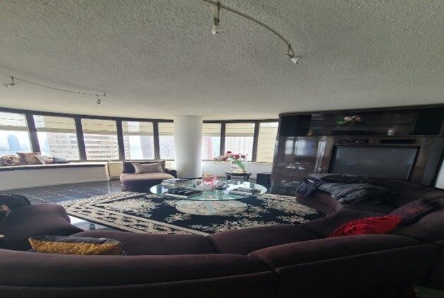 Spacious living room with large curved brown sectional sofa around a glass coffee table on a patterned rug, large windows with city views, and built-in dark wood entertainment center.