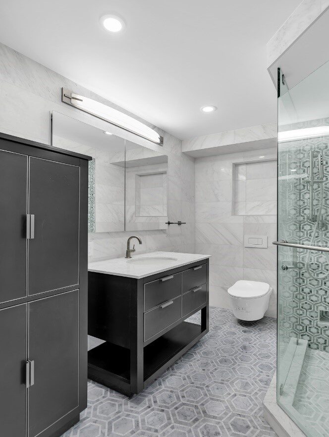 Modern bathroom with a black vanity, white countertop, wall-mounted toilet, and glass shower with patterned tile.