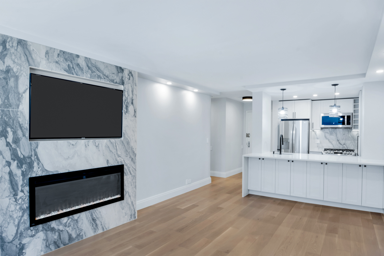 Modern open-concept living space with light wood floors, white kitchen cabinets, marble accent wall with mounted TV and electric fireplace.