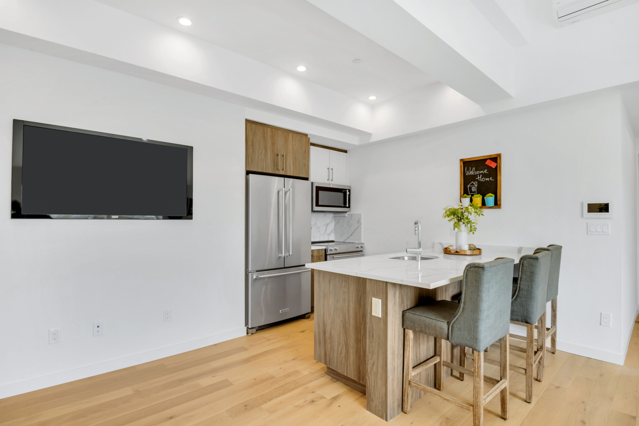 Modern kitchen with a wooden island, three gray upholstered bar stools, stainless steel refrigerator, mounted TV, and a small chalkboard saying 'Welcome Home'.