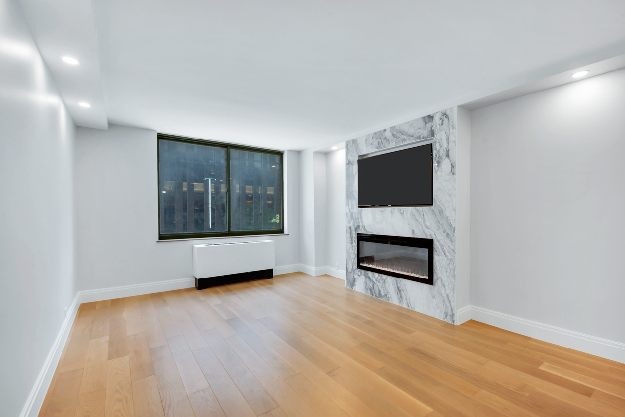 Empty modern living room with wooden floor, large window, wall-mounted TV, and built-in electric fireplace with marble surround.
