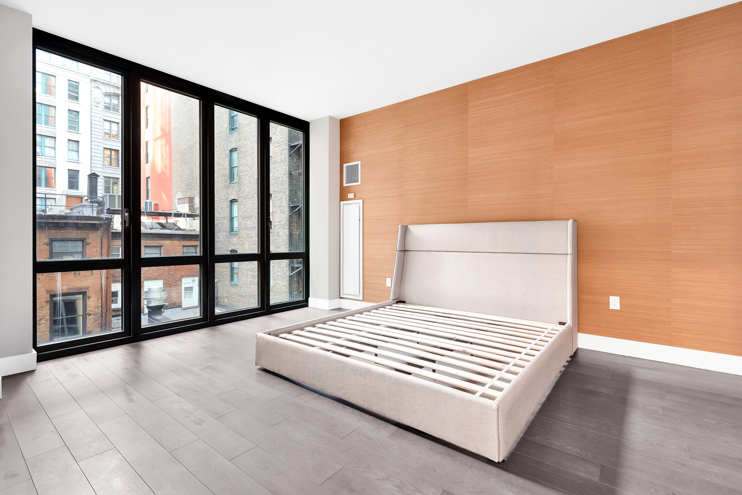 Modern bedroom with large floor-to-ceiling windows overlooking city buildings and a beige upholstered bed frame without mattress.