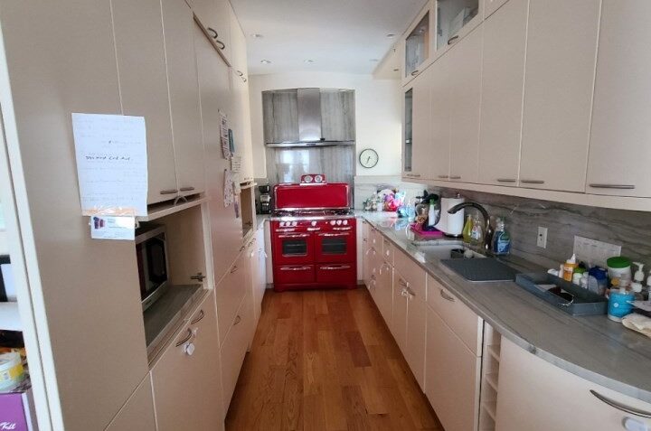 Narrow kitchen with light beige cabinets, wooden floor, gray countertops, and a red vintage-style stove at the end.