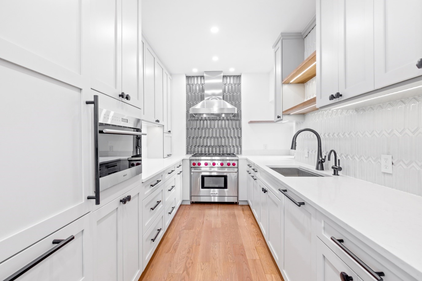 Modern white kitchen with stainless steel stove, black handles, wooden floor, and geometric tile backsplash.