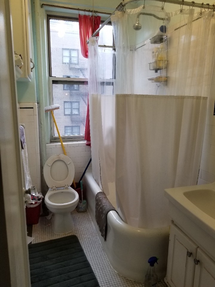 Small bathroom with white tiled floor, toilet with open lid, bathtub with a white shower curtain, and a window with red curtains.