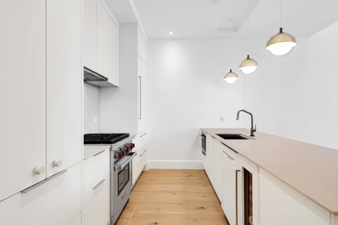 Modern minimalist kitchen with white cabinets, stainless steel oven and stovetop, wooden floor, beige countertop, black sink, and three pendant lights.