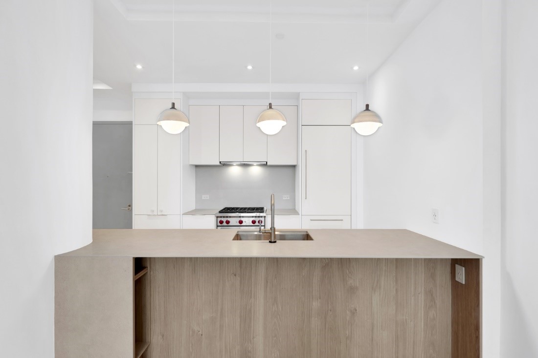 Modern white kitchen with beige countertop island, stainless steel sink, three hanging pendant lights, and built-in stove and refrigerator.