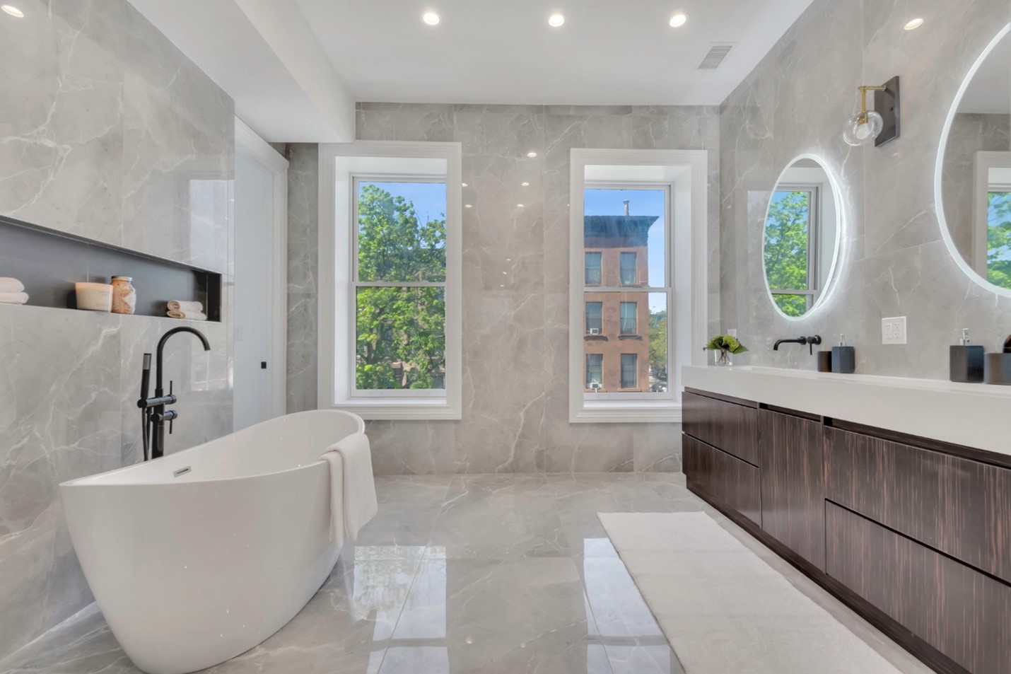 Modern bathroom with freestanding white bathtub, large gray marble tiles, double vanity with dark wood cabinets, and two round illuminated mirrors.