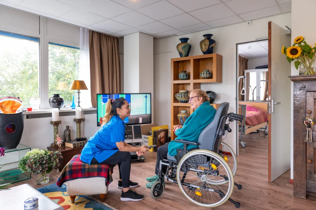 A caregiver in blue scrubs kneeling and smiling while talking to an elderly woman in a wheelchair in a bright, decorated living room.
