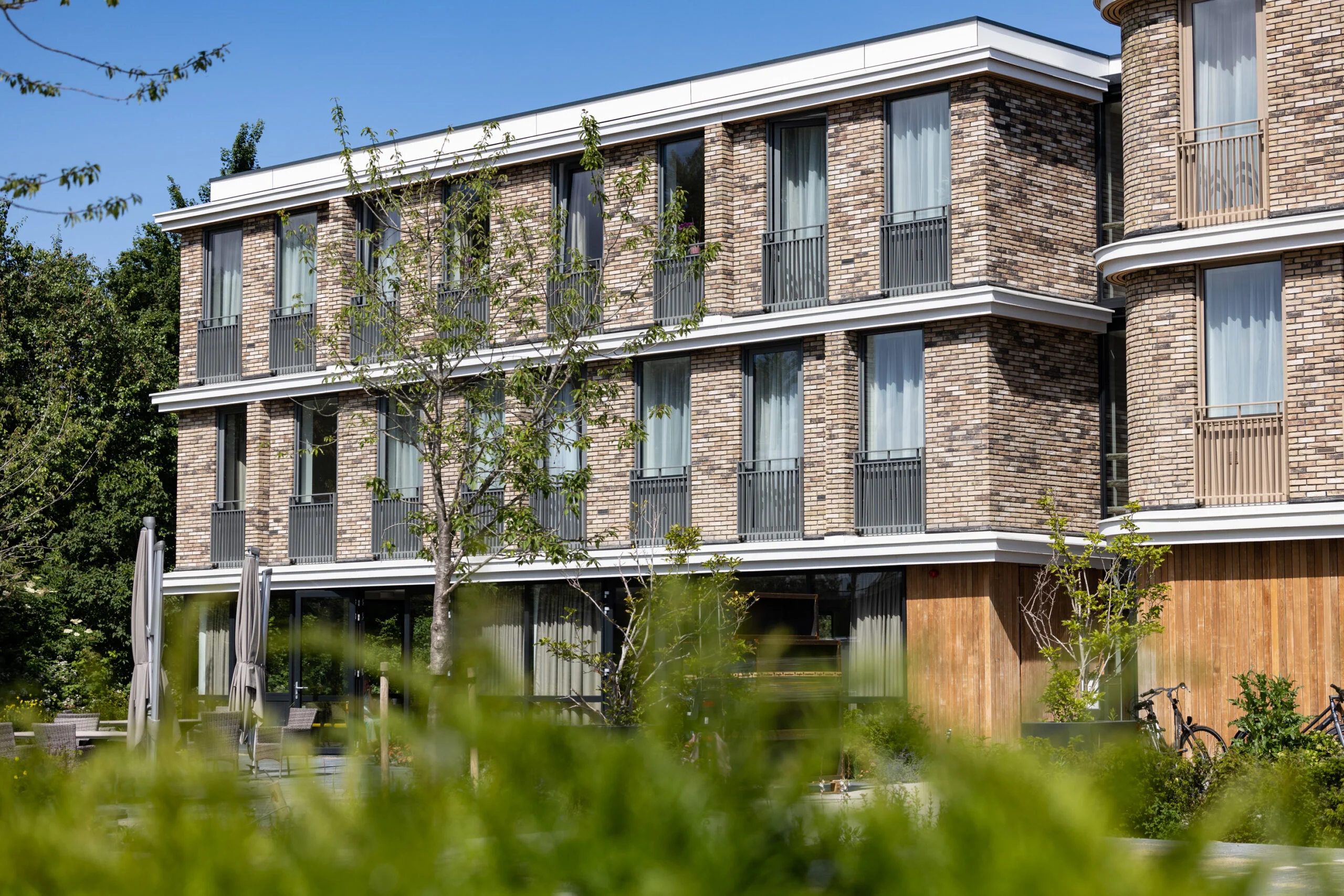 Modern three-story brick building with large windows, greenery, outdoor seating, and bicycles parked nearby under a clear blue sky.