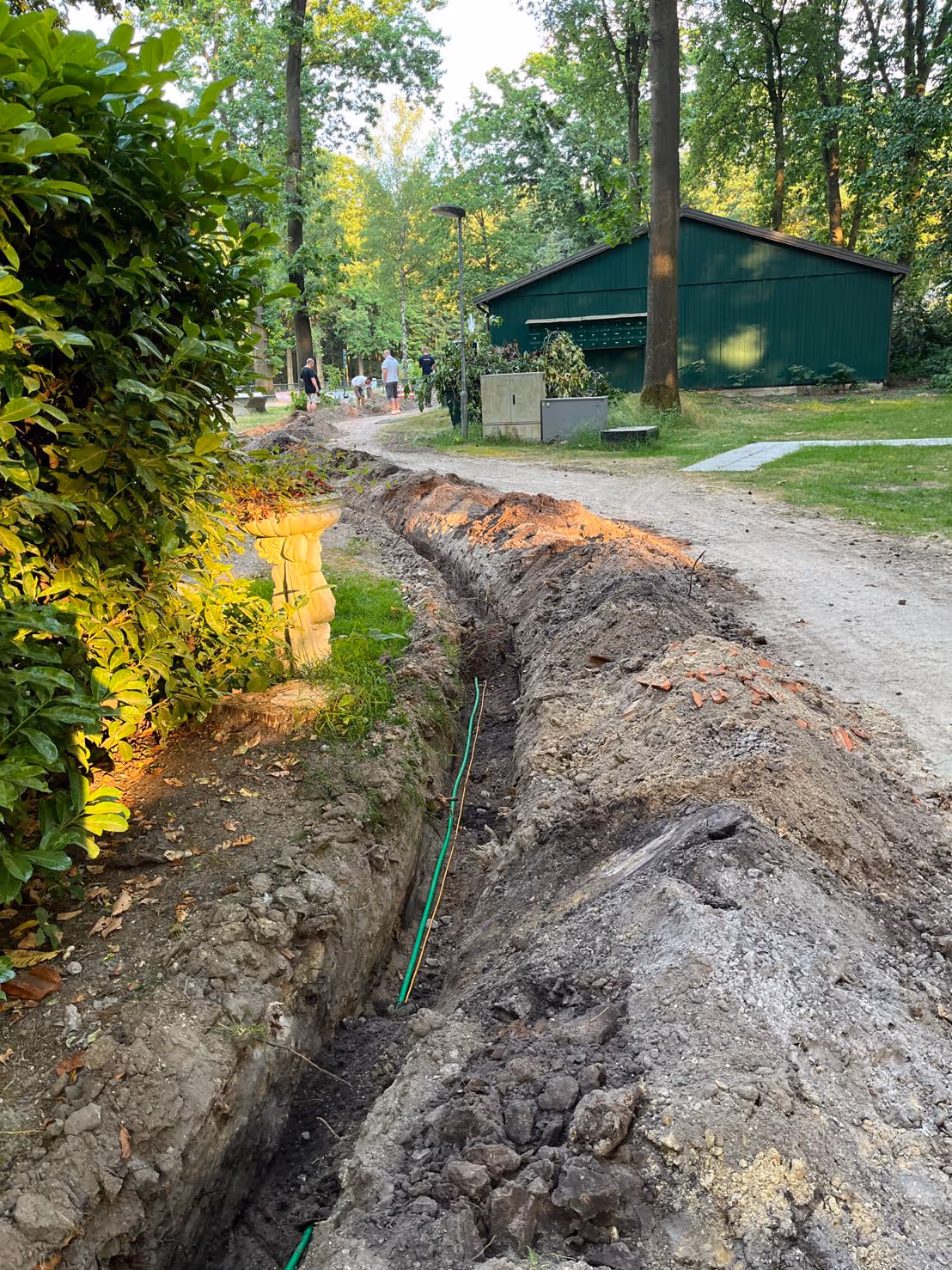 Long trench dug along a dirt path in a green park with people working in the background and a green shed nearby.