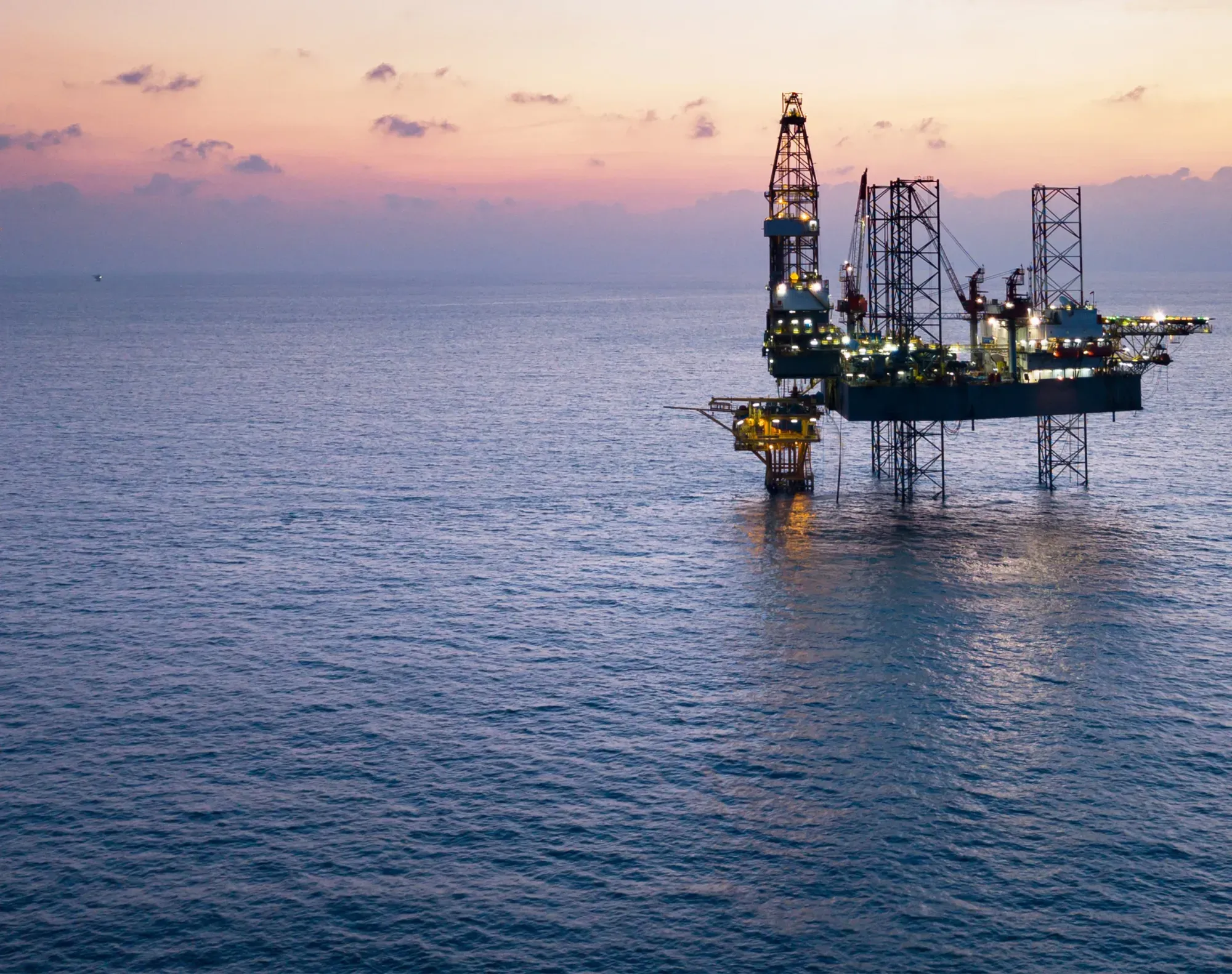 Illuminated offshore oil rig standing in calm ocean waters at sunset.