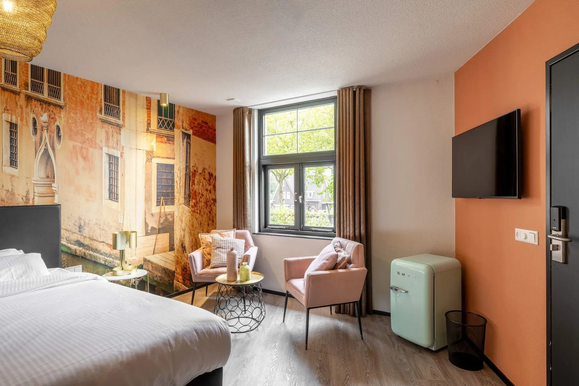 Cozy hotel room with a large bed, two pink armchairs and a round table near a window, a wall-mounted TV, and a small retro mint green refrigerator.