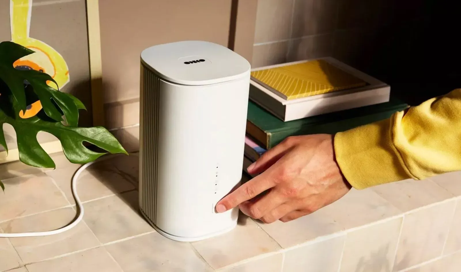 Hand pressing a button on a white Zyxel router placed on a tiled surface next to books and a plant leaf.