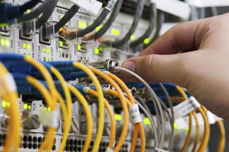 Hand connecting a coaxial cable to a network switch with multiple colored cables and glowing indicator lights.