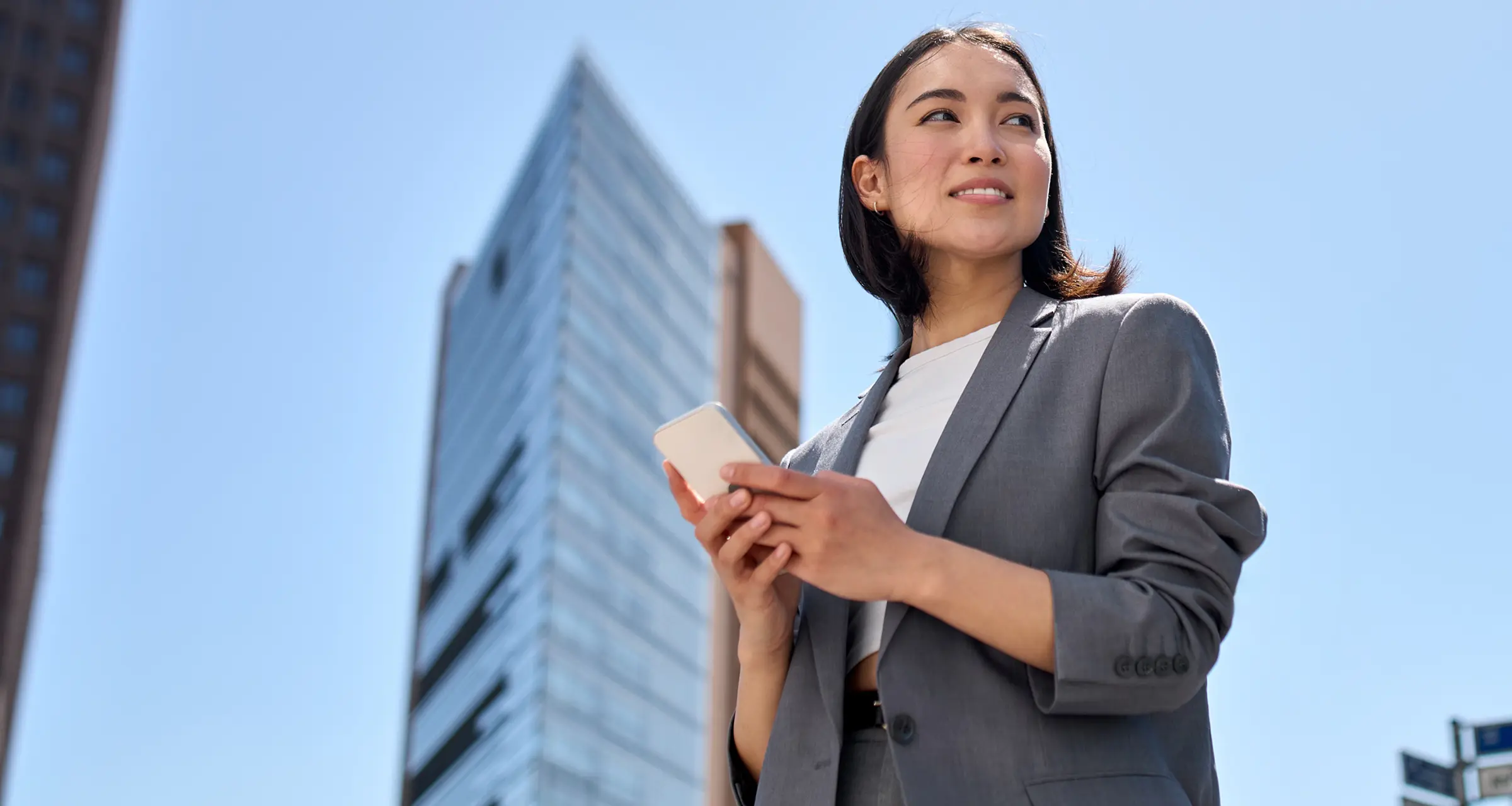 Confident woman in a gray business suit holding a smartphone with a modern skyscraper in the background.