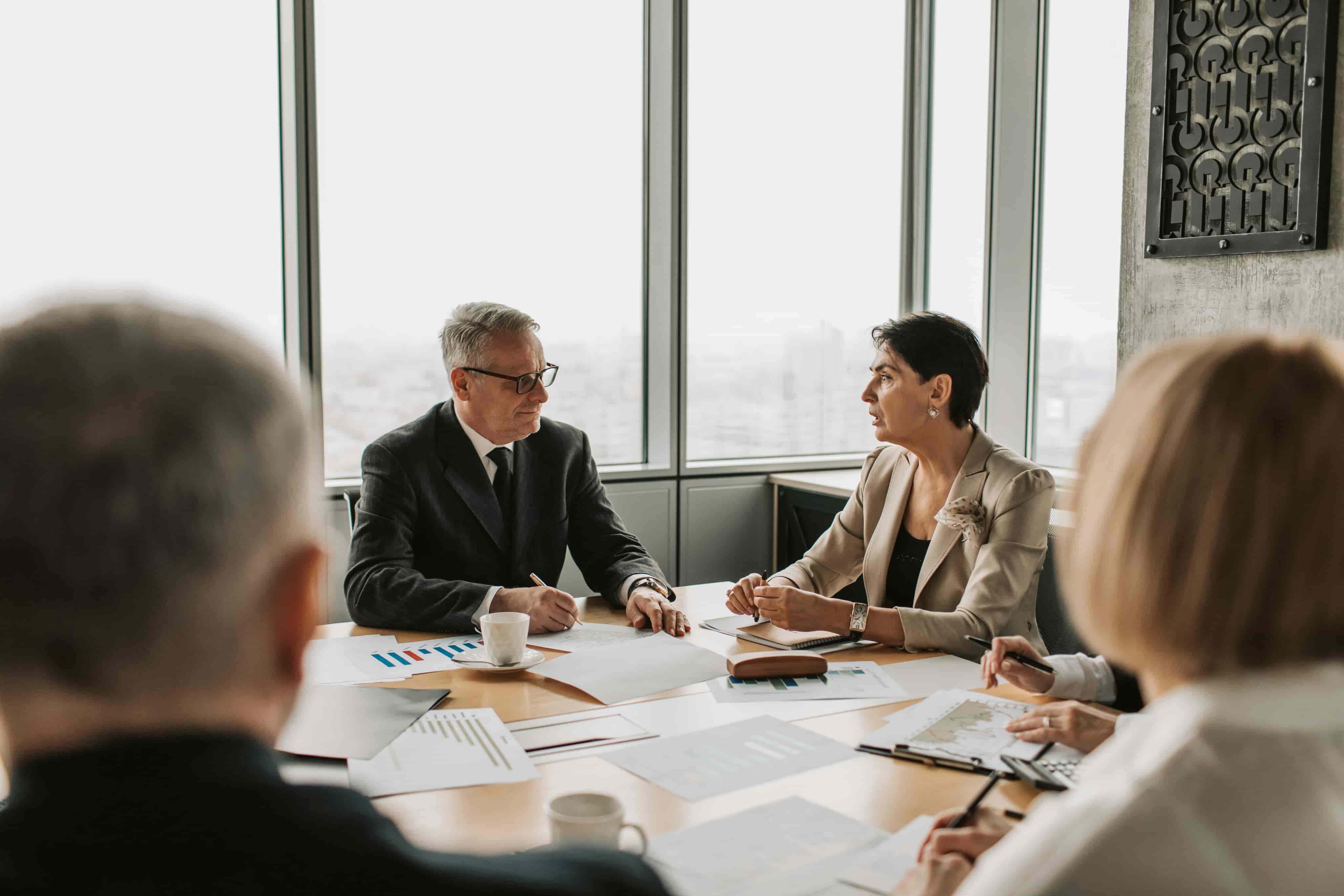 Four professionals in a business meeting around a table with charts and documents, two are seen clearly discussing business.