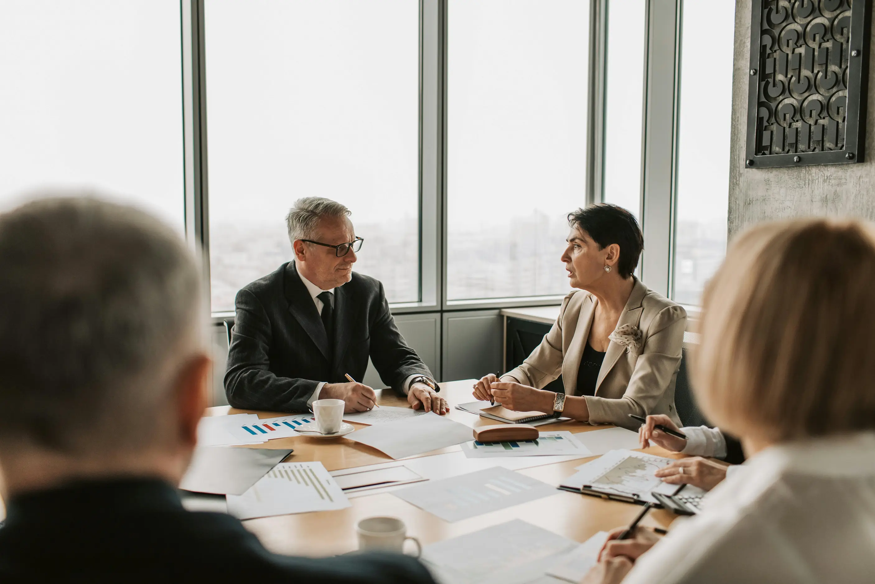 Four professionals in a business meeting around a table with charts and documents, two are seen clearly discussing business.
