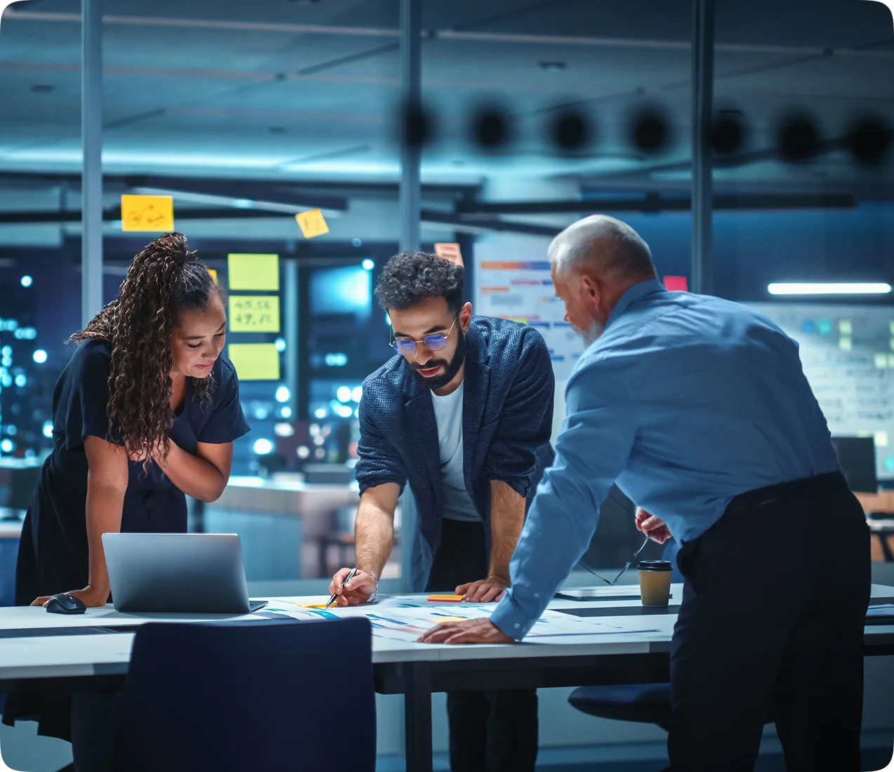 Three colleagues in an office at night discussing documents on a table with a laptop and sticky notes on the glass wall behind them.