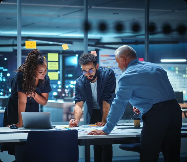 Three colleagues in an office at night discussing documents on a table with a laptop and sticky notes on the glass wall behind them.