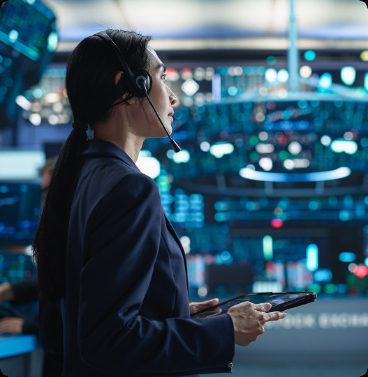 Woman wearing headset and holding tablet, standing in front of multiple digital screens in a control room.