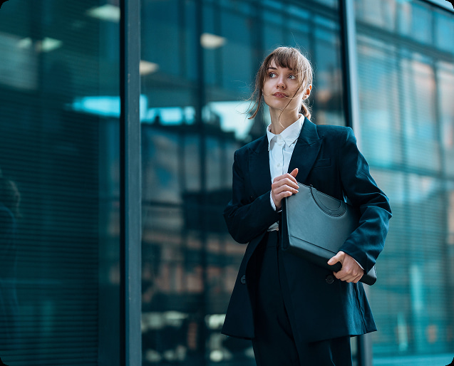 Young woman in a dark suit holding a black portfolio standing in front of a modern glass building.