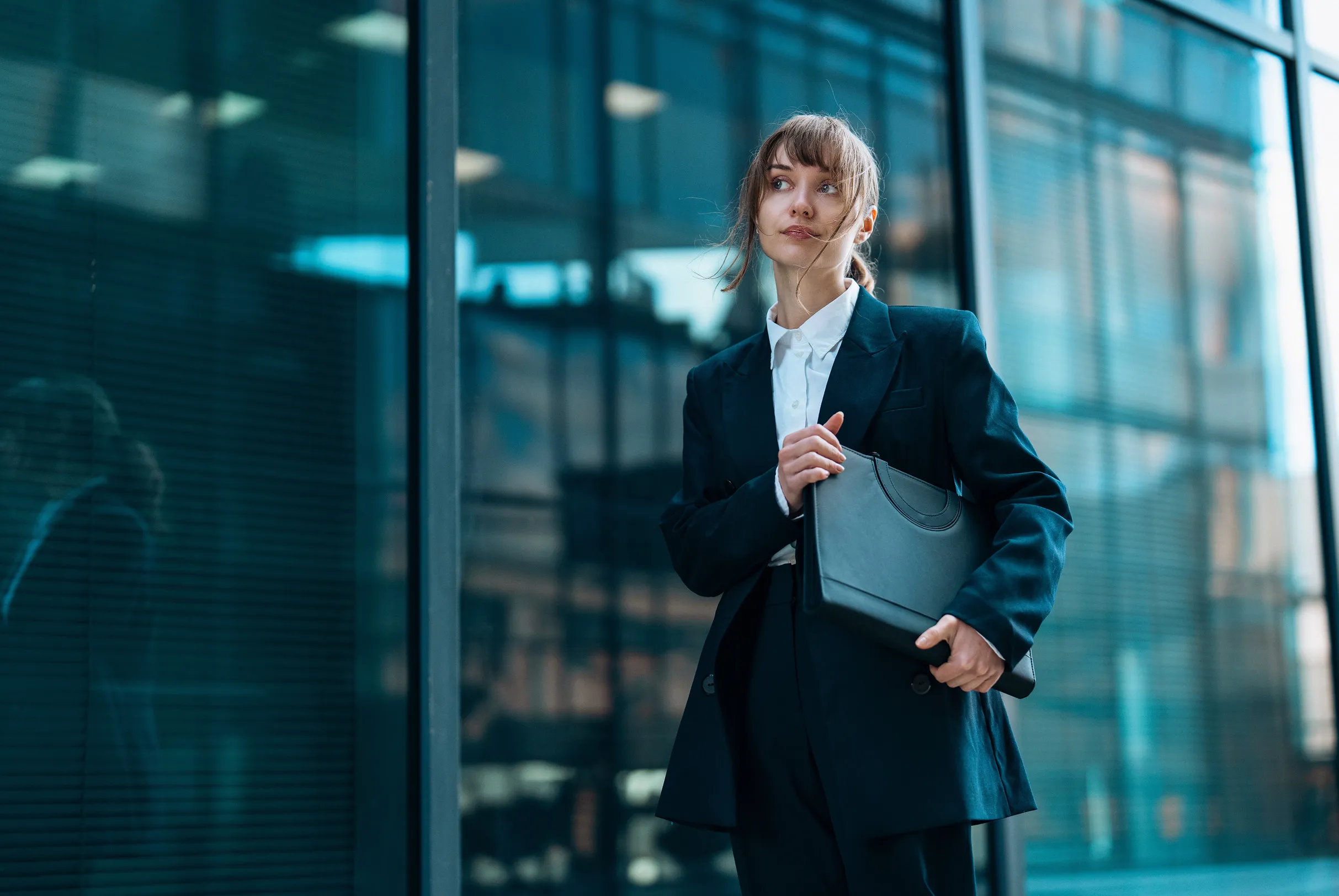 Young woman in a dark suit holding a black portfolio standing in front of a modern glass building.