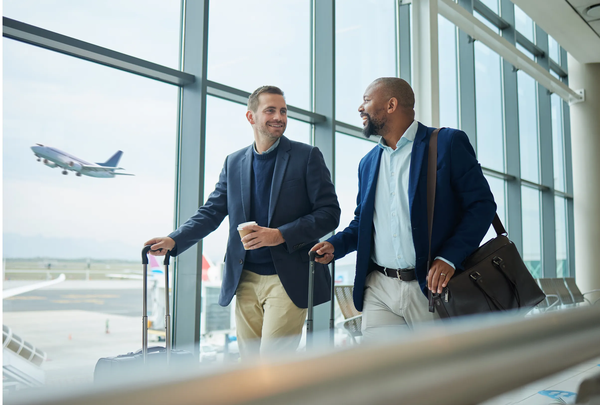 Two businessmen walking together at an airport terminal pulling suitcases and chatting.