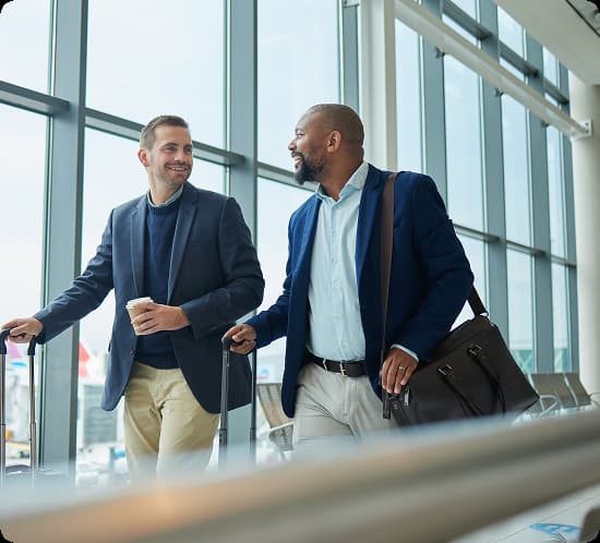 Two businessmen walking together at an airport terminal pulling suitcases and chatting.