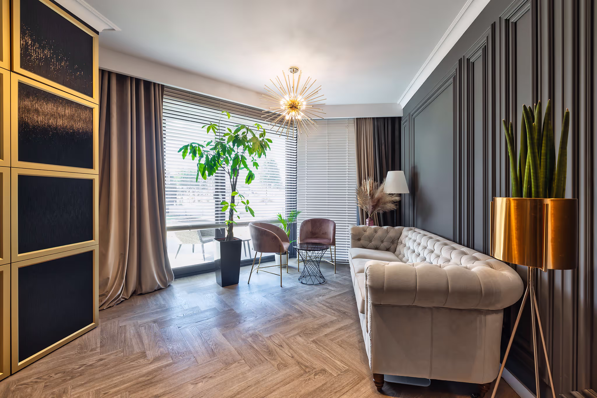 Nowoczesny salon z jasnoszarą sofą, dwoma fioletowymi fotelami, dużą rośliną przy oknie i stojącą lampą z złotym abażurem.