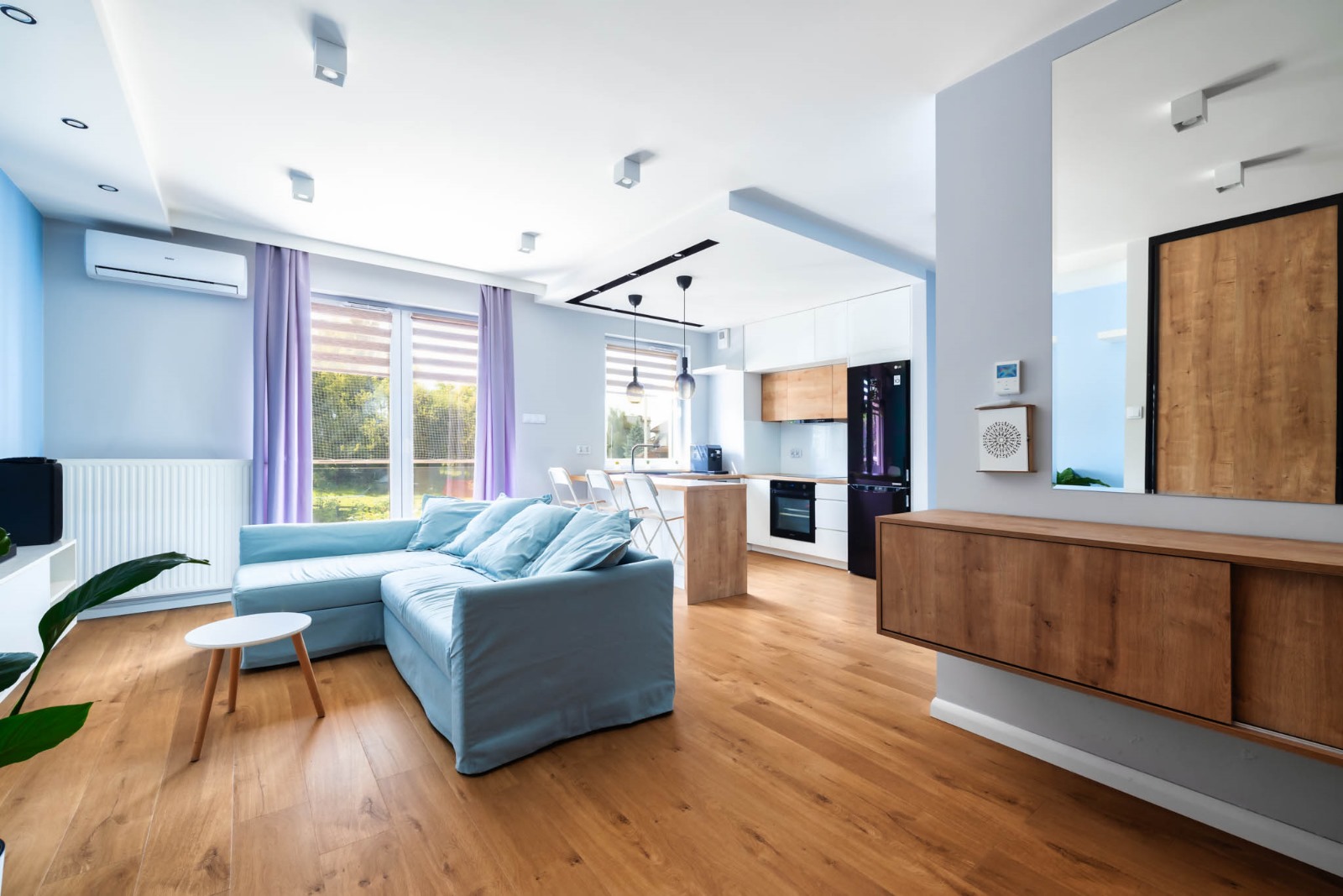 Nowoczesny salon z jasnoniebieską sofą, drewnianą podłogą i kuchnią z wyspą oraz trzema krzesłami barowymi.