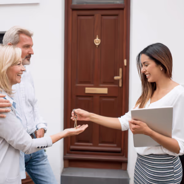 Kobieta przekazuje klucze do domu uśmiechającej się parze stojącej przed drewnianymi drzwiami.